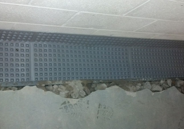 Damaged interior wall with exposed rubble beneath a gray patterned panel and white ceiling above